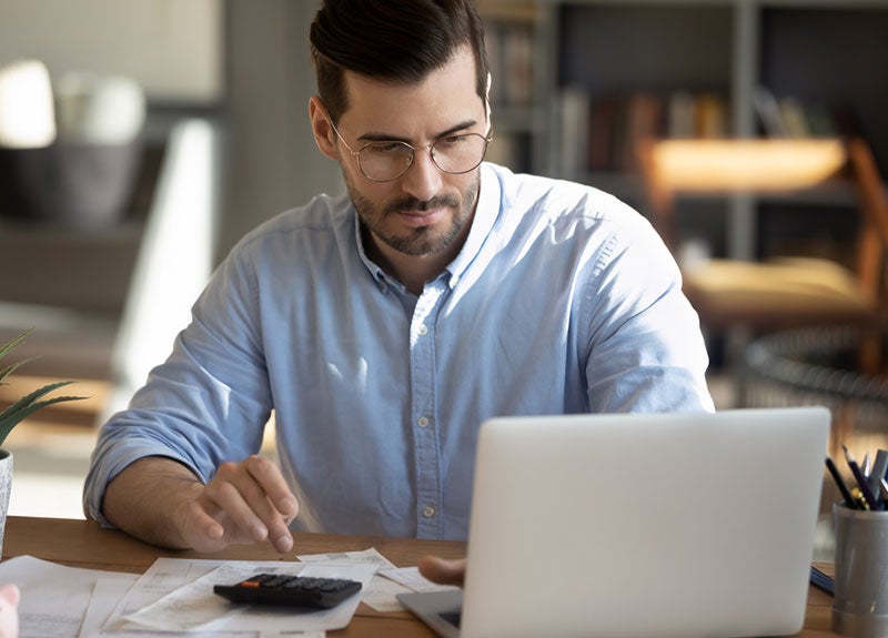 Focused man in glasses calculating finances with a calculator, receipts, and laptop on a wooden desk.