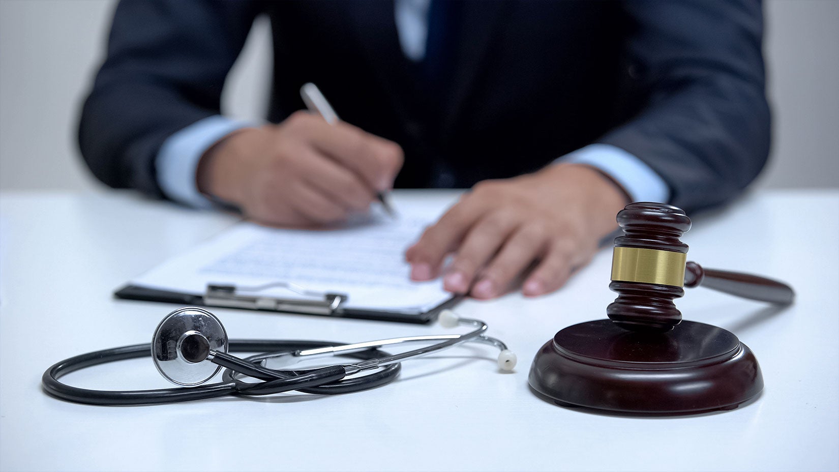 A person in a suit writes on a clipboard next to a stethoscope and a judge's gavel on a white table.