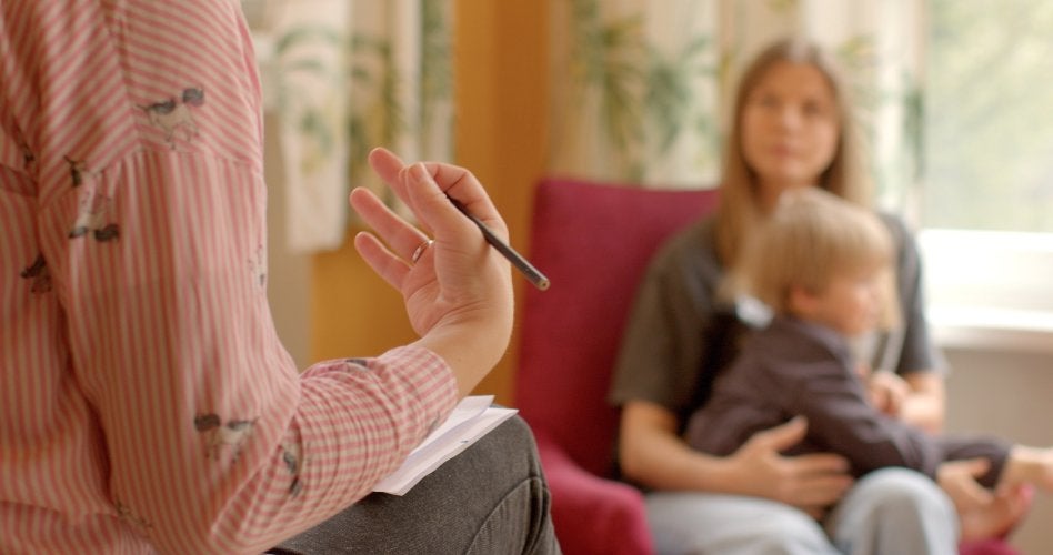 A therapist’s hand holding a pen while talking to a mother holding her child in a blurry background.