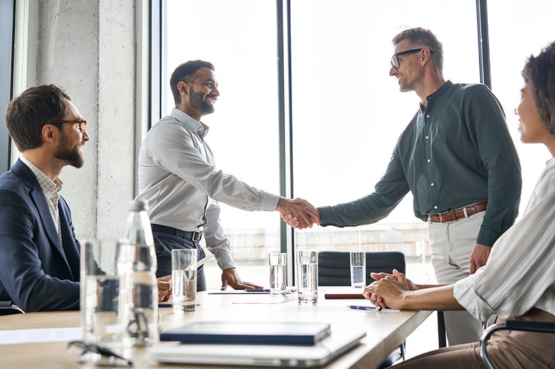 Two men in an office boardroom shake hands while a bearded man and a woman look on.