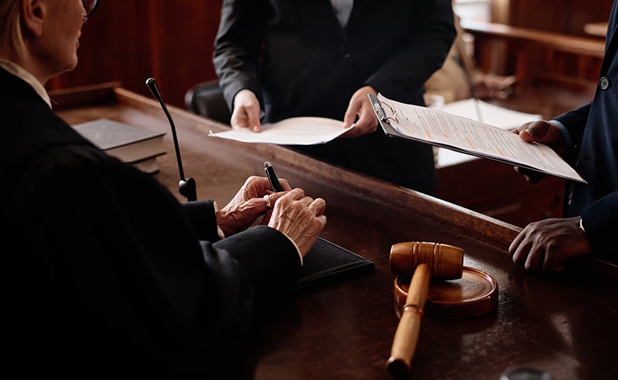 A judge signs documents at a courthouse bench with a gavel next to them, while lawyers exchange papers.