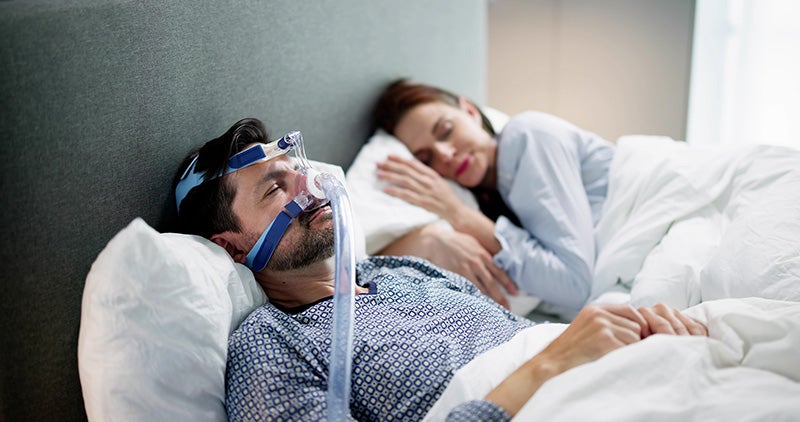 A man wearing a sleep apnea mask sleeps in bed next to a sleeping woman.