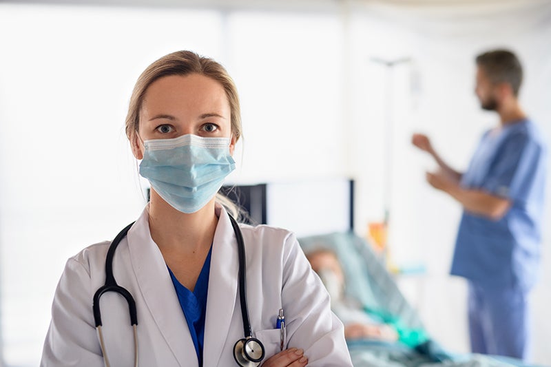 Female doctor in white coat and surgical mask, with blurred colleagues and patient in a hospital room.