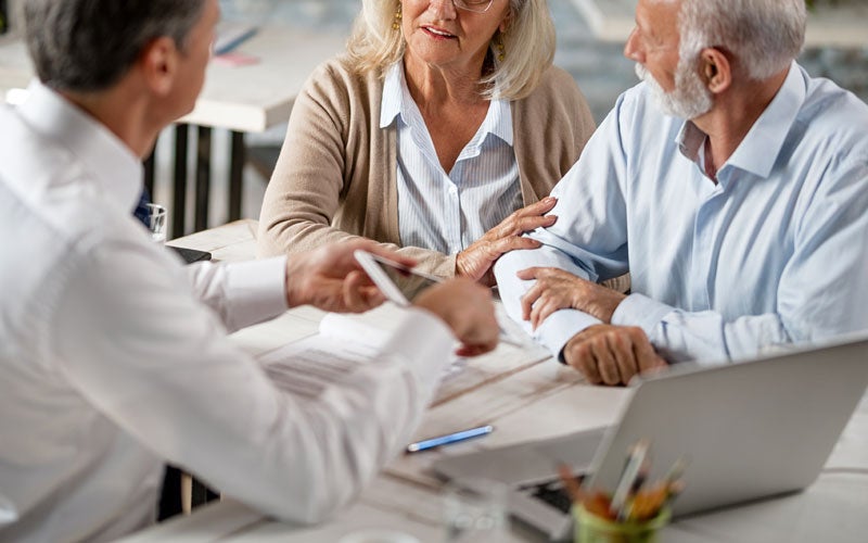 A financial advisor explains information on a tablet to an older couple sitting at a table with a laptop.
