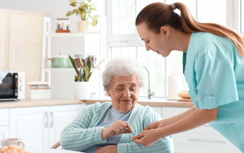 A young caretaker in scrubs helps an elderly woman take her medication in a bright kitchen.