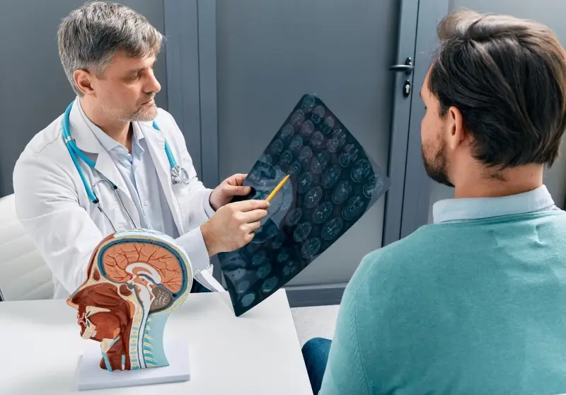 A doctor in a white coat shows a brain scan to a patient while pointing with a pencil.