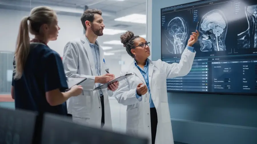 Three medical professionals, two men and one woman, examine brain MRI scans on a large screen in a medical facility.