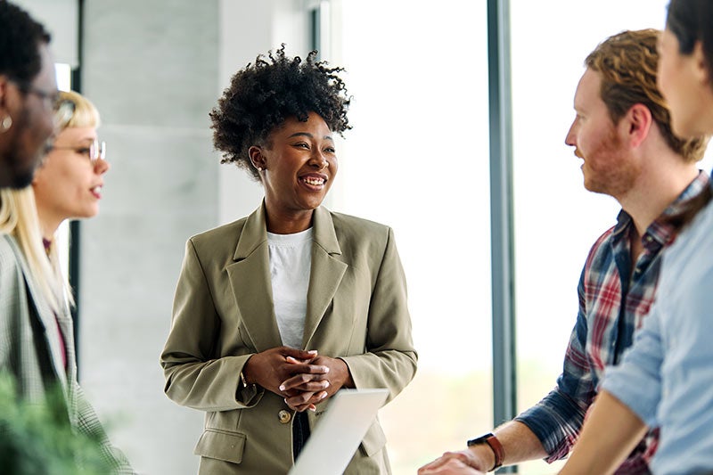 A smiling Black woman in a blazer speaks with three diverse colleagues in a bright office.